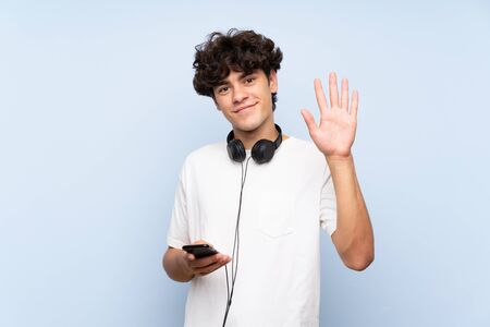 Young man listening music with a mobile over isolated blue wall saluting with hand with happy expressionの写真素材
