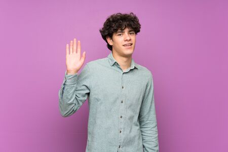 Young man over isolated purple wall saluting with hand with happy expressionの写真素材