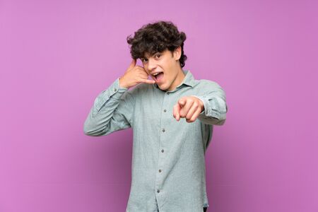 Young man over isolated purple wall making phone gesture and pointing frontの写真素材
