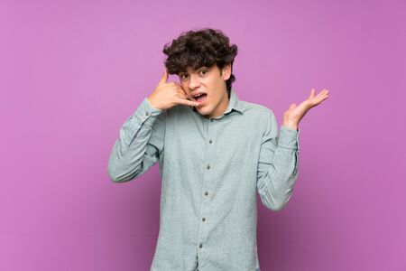 Young man over isolated purple wall making phone gesture and doubtingの写真素材