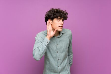 Young man over isolated purple wall listening to something by putting hand on the earの写真素材