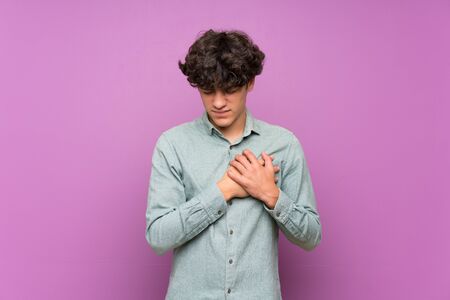 Young man over isolated purple wall having a pain in the heartの写真素材
