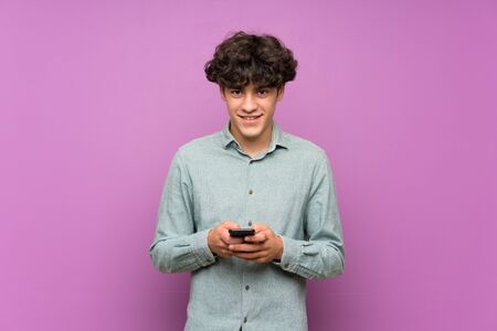 Young man over isolated purple wall sending a message with the mobileの写真素材