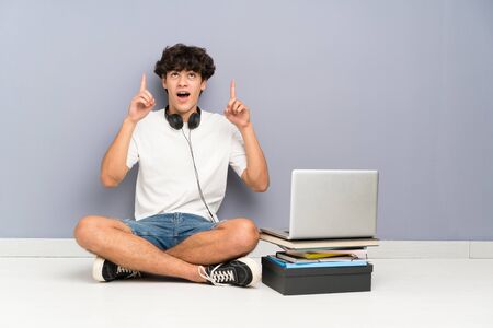 Young man with his laptop sitting one the floor pointing with the index finger a great ideaの写真素材