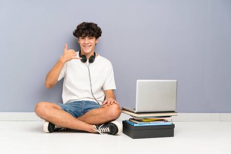 Young man with his laptop sitting one the floor making phone gestureの写真素材