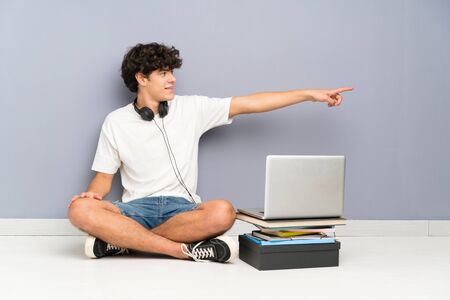 Young man with his laptop sitting one the floor pointing finger to the sideの写真素材