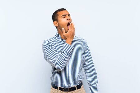 Young handsome brunette man over isolated blue background yawning and covering wide open mouth with handの写真素材