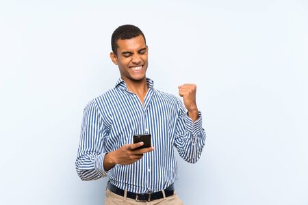 Young handsome brunette man over isolated blue background with phone in victory positionの写真素材