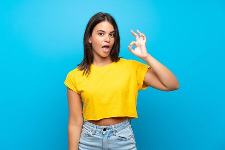 Young girl over isolated blue background surprised and showing ok signの写真素材