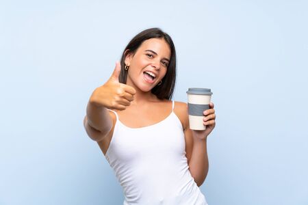 Young woman holding a take away coffee over isolated blue background with thumbs up because something good has happenedの写真素材