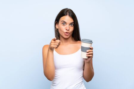 Young woman holding a take away coffee over isolated blue background surprised and pointing frontの写真素材