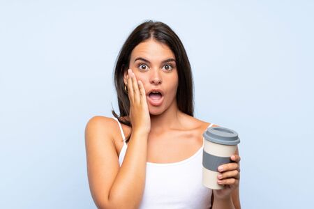 Young woman holding a take away coffee over isolated blue background with surprise and shocked facial expressionの写真素材