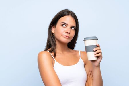 Young woman holding a take away coffee over isolated blue background making doubts gesture while lifting the shouldersの写真素材