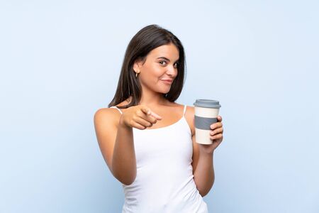 Young woman holding a take away coffee over isolated blue background points finger at you with a confident expressionの写真素材