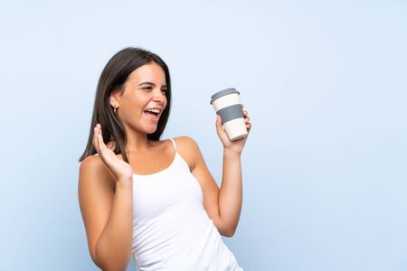 Young woman holding a take away coffee over isolated blue background with surprise facial expressionの写真素材