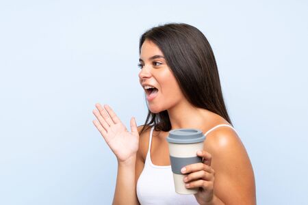 Young woman holding a take away coffee over isolated blue background shouting with mouth wide openの写真素材