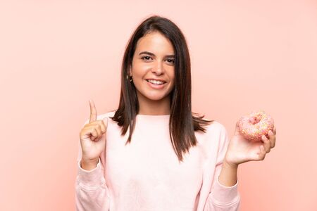 Young girl holding a donut over isolated pink background pointing up a great ideaの写真素材