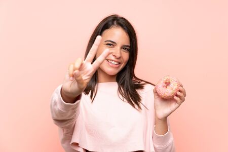 Young girl holding a donut over isolated pink background smiling and showing victory signの写真素材