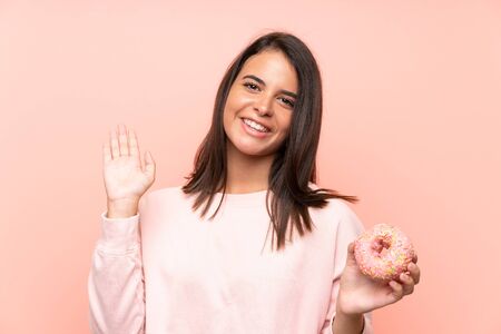 Young girl holding a donut over isolated pink background saluting with hand with happy expressionの写真素材