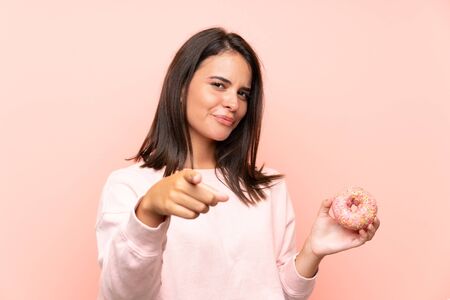 Young girl holding a donut over isolated pink background points finger at you with a confident expressionの写真素材
