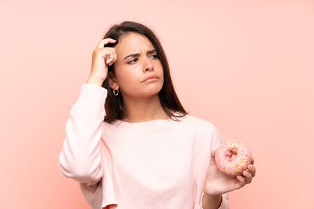 Young girl holding a donut over isolated pink background having doubts and with confuse face expressionの写真素材