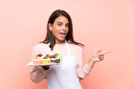 Young girl holding lots of different mini cakes over isolated background surprised and pointing finger to the sideの写真素材