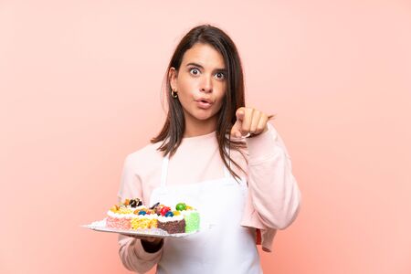 Young girl holding lots of different mini cakes over isolated background surprised and pointing frontの写真素材