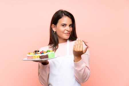 Young girl holding lots of different mini cakes over isolated background inviting to come with hand. Happy that you cameの写真素材