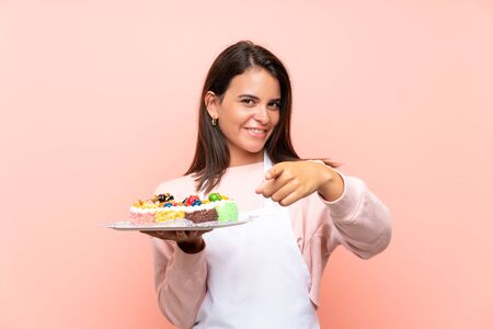 Young girl holding lots of different mini cakes over isolated background points finger at you with a confident expressionの写真素材