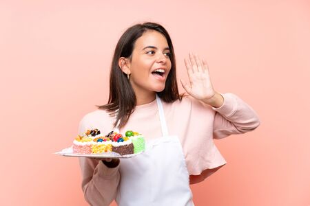 Young girl holding lots of different mini cakes over isolated background shouting with mouth wide openの写真素材