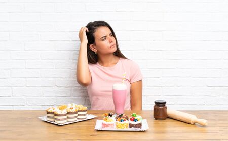 Young girl with lots of different mini cakes having doubts and with confuse face expressionの写真素材