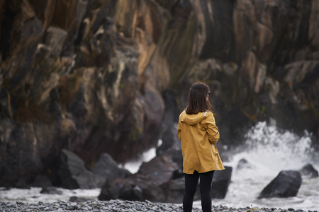 Woman on a yellow rain jacket looking to the sea with rocks on the background, in Madeiraの写真素材