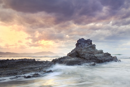 Seascape showing waves breaking against a rock on the coast with a cloudy stormy sky and the sun appearing in the backgroundの写真素材