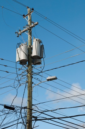 messy old electricity post by the road with power line cables, transformers and phone lines (blue sky background)の写真素材