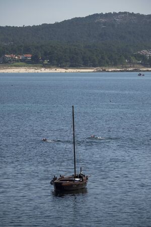 O GROVE, SPAIN - AUGUST 12, 2019: Wooden boat anchored on the coast and two swimmers behind it. Quaint traditional and ancient boat on the Galician coastのeditorial素材