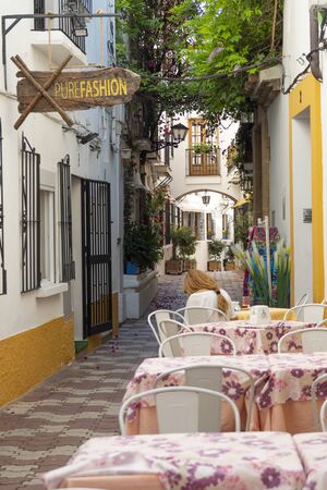 MARBELLA, SPAIN - JUNE 21, 2019: Street of the historic center of Marbella, Spain. Street view with restaurant tables in the foreground and a blonde woman sittingのeditorial素材