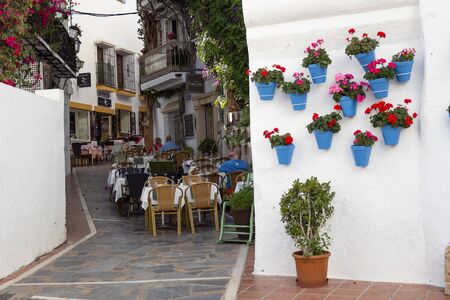 MARBELLA, SPAIN - JUNE 21, 2019: Picturesque typical street of Marbella, Spain. With beautiful blue pots with red geraniums and the tables of a restaurant.のeditorial素材