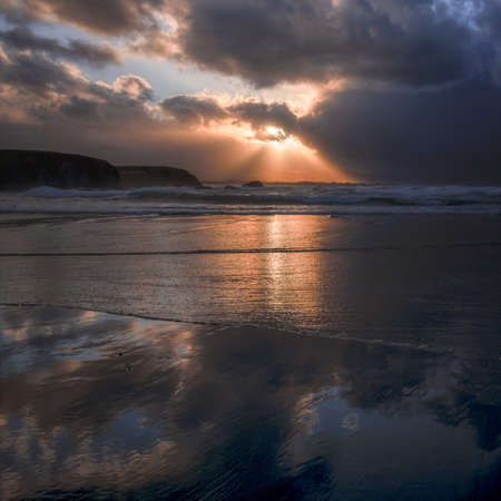 Golden rays of the setting sun between the clouds of a sky reflected on the wet shore of a beachの写真素材