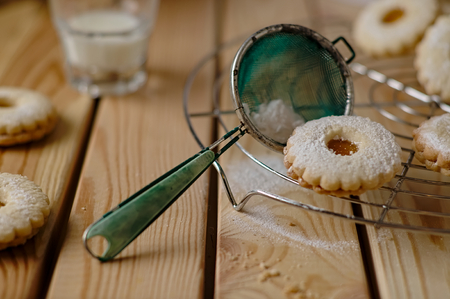 Sugared Linz biscuit with peach marmalade on a cooling grate on top of a wooden table with crumbs with a glass of milk in the backgroundの写真素材