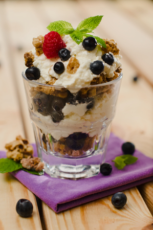 Sweet fresh cottage-cheese and oat flake granola dessert topped with blueberries, raspberry and a mint leaf on a violet napkin with a wooden table background.の写真素材