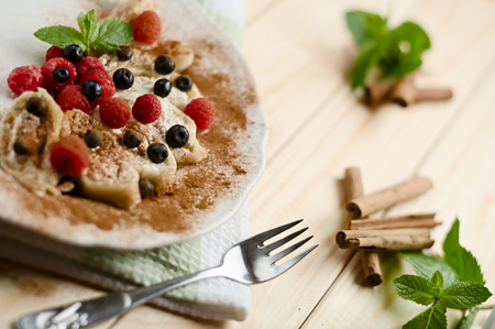 Sweet homemade dessert fresh cottage cheese dumpling rolls with raspberries, blueberries, and powdered cinnamon on a wood table decorated with a kitchen cloth, fork, cinnamon sticks and mint leavesの写真素材