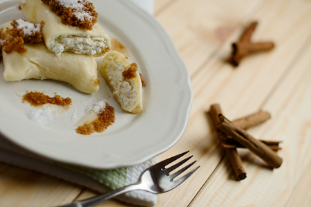 Delicious homemade sweet fresh cottage cheese dumpling rolls with pan fried breadcrumbs and granulated sugar and cinnamon on a wood table decorated with a kitchen cloth, fork, and cinnamon sticksの写真素材