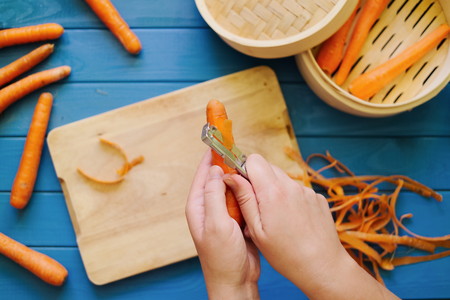 female hands peeling an orange carrot on blue background. Concept of healthy food preparation. Peeled carrots in steamer. Hands in frame. Top down view. Flat lay.の写真素材