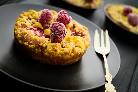 Sweet homebaked raspberry jam crumble tart with frozen raspberries on black plate on black wood boards with gold fork. Selective focus on the front raspberry. Concept of home baking.の写真素材