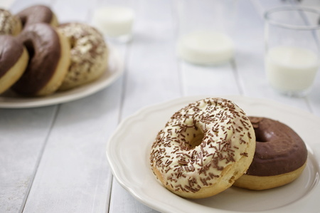 Sweet homemade dessert doughnuts with white and milk chocolate in rustic setting on white wood boards and glasses of milk and milk bottle. Selective focus on front doughnut.の写真素材