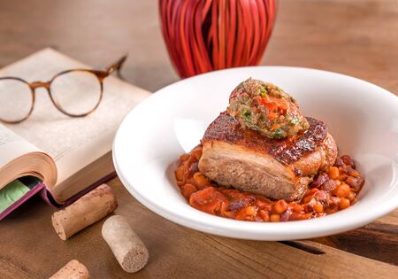 Pork belly with bean stew and mashed vegetables, served in a white plate, decorated with book and glasses on a wooden table.の写真素材