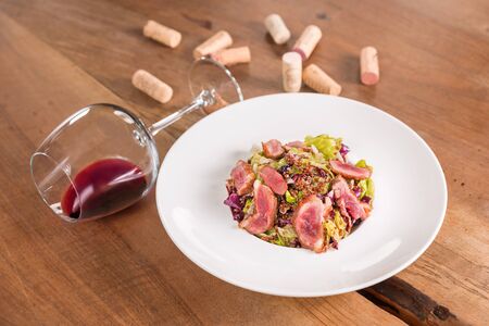 Duck breast salad with glass of red wine, served on a white plate, decorated with corks.の写真素材