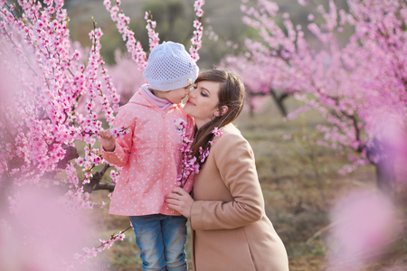 Cute beautiful stylish dressed brunette girl with mother mom standing on a field of spring young peach tree with pink flowers.Lady dressed in rain clothes and blue hat rubber boots spring seasonの写真素材