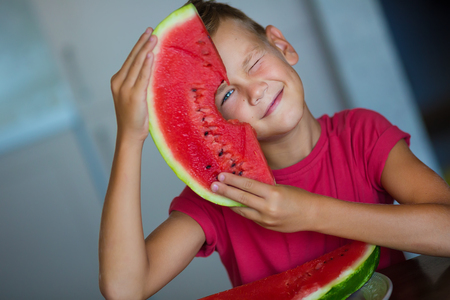 Cute kid enjoy summer juicy berry fruit watermelon Handsome blue eyes Boy wearing red t-shirt with slice of watermelon isolated on home studio kitchenの写真素材