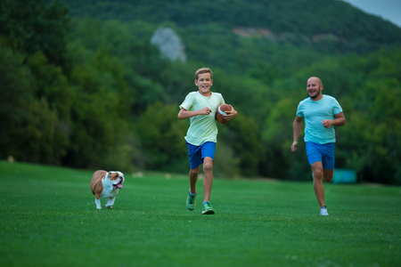 Father and son are running in park with dog english bulldog. Father's Day. Father with son and dog on a walk in the city park on sunny summer day. Family walking with dog in park.の写真素材